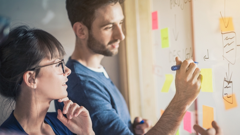 Decorative image of two adults brainstorming. The adult man is making notes on a board and the adult woman is deep in thought beside him