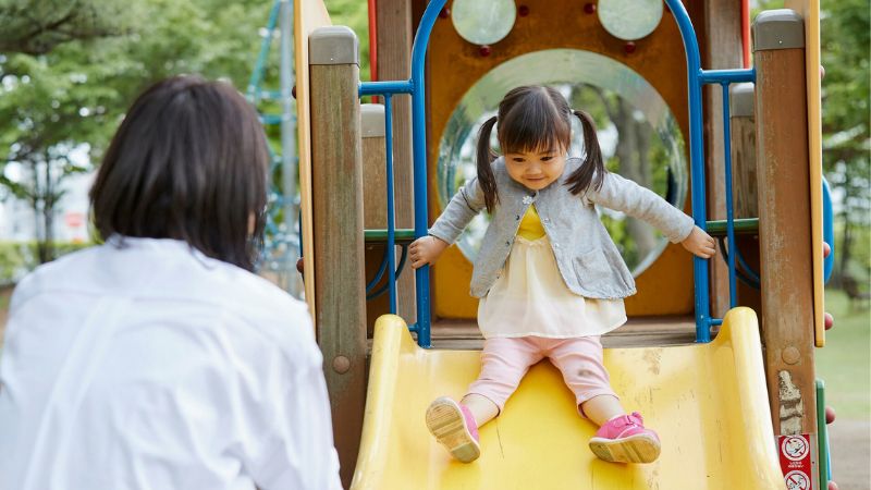 Decorative image of a little girl sliding down a slide at the playground while her mother watches