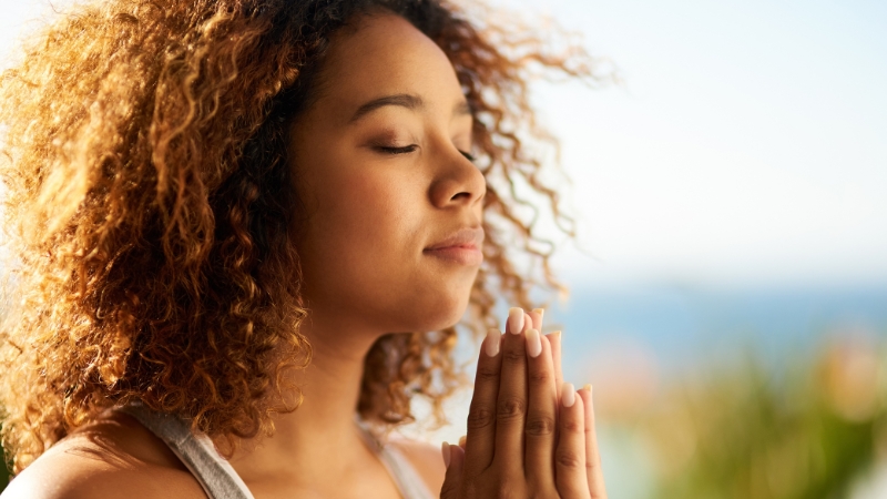 Decorative image of a black woman with her hands clasped, praying or meditating quietly while the sun shines down on her from behind