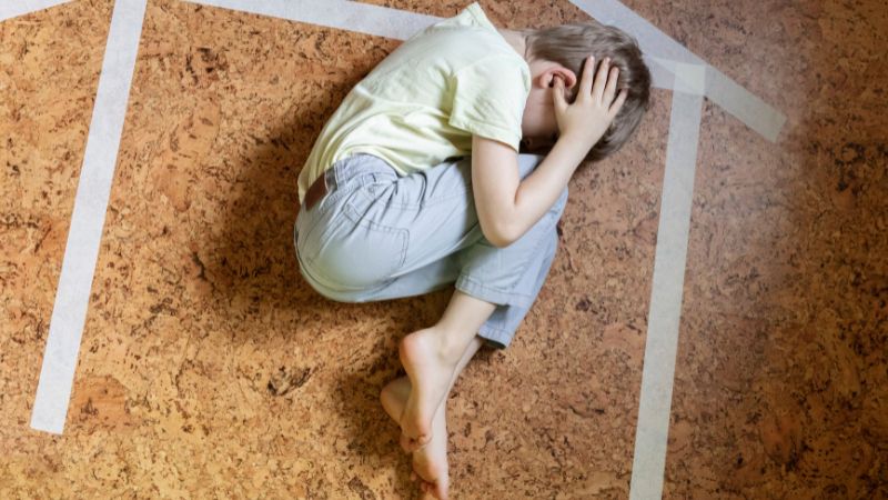 Decorative image of a child curled up with his head in his hands on the floor, where a picture of a house has been drawn with paint