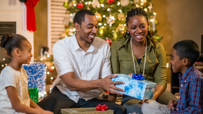 Family sharing Christmas gifts for the holidays. The father is handing a present over to his son while his daughter and wife watch.