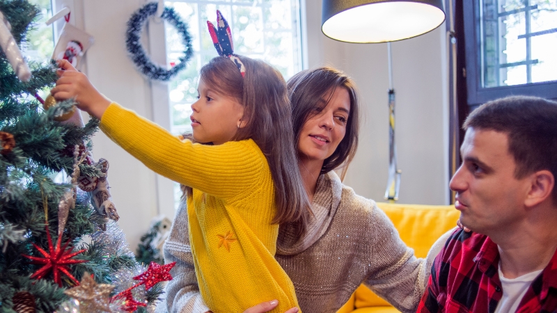 Decorative image of a child placing a decoration on a Christmas tree while her parents watch