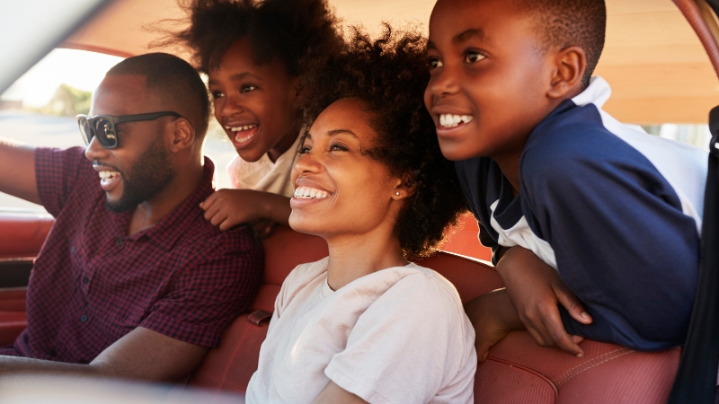 Decorative image of a family of four. The parents are seated at the front of their car, and their kids are peering from the backseat.