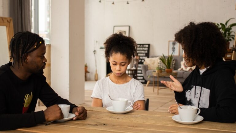 Decorative image of co parents seated at the table with their child. The mother is in the middle of explaining something. The father is listening. The child has her head lowered and arms crossed.