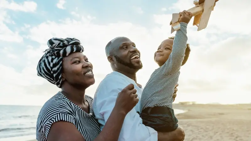 Decorative image of a black family of three. Both parents wear smiles on their faces as their son holds a wooden plane toy against the sky